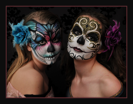 two women in full face paint that are skull themed and highly stylized. One with blue and pink highlights they other in black and gold